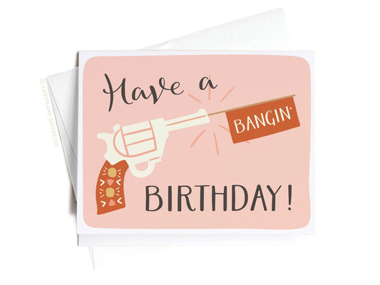 Hand holding a birthday card with a gun graphic and text on a pink background saying have a bangin birthday