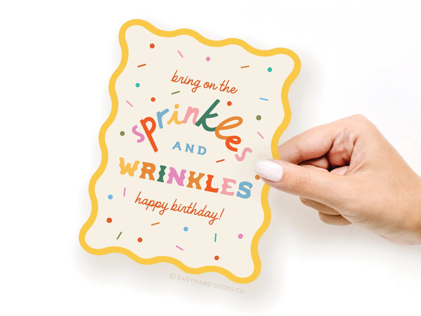 Hand holding a colorful birthday card with text 'bring on the sprinkles and wrinkles happy birthday' on a white background