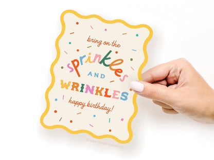 Hand holding a colorful birthday card with text 'bring on the sprinkles and wrinkles happy birthday' on a white background