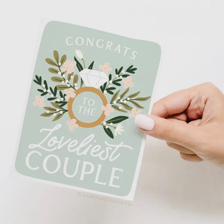 Hand holding a card with 'Congrats to the Loveliest Couple' text and floral design on a light background