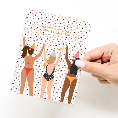 Hand holding a birthday card with illustrations of women in swimsuits and party hats.