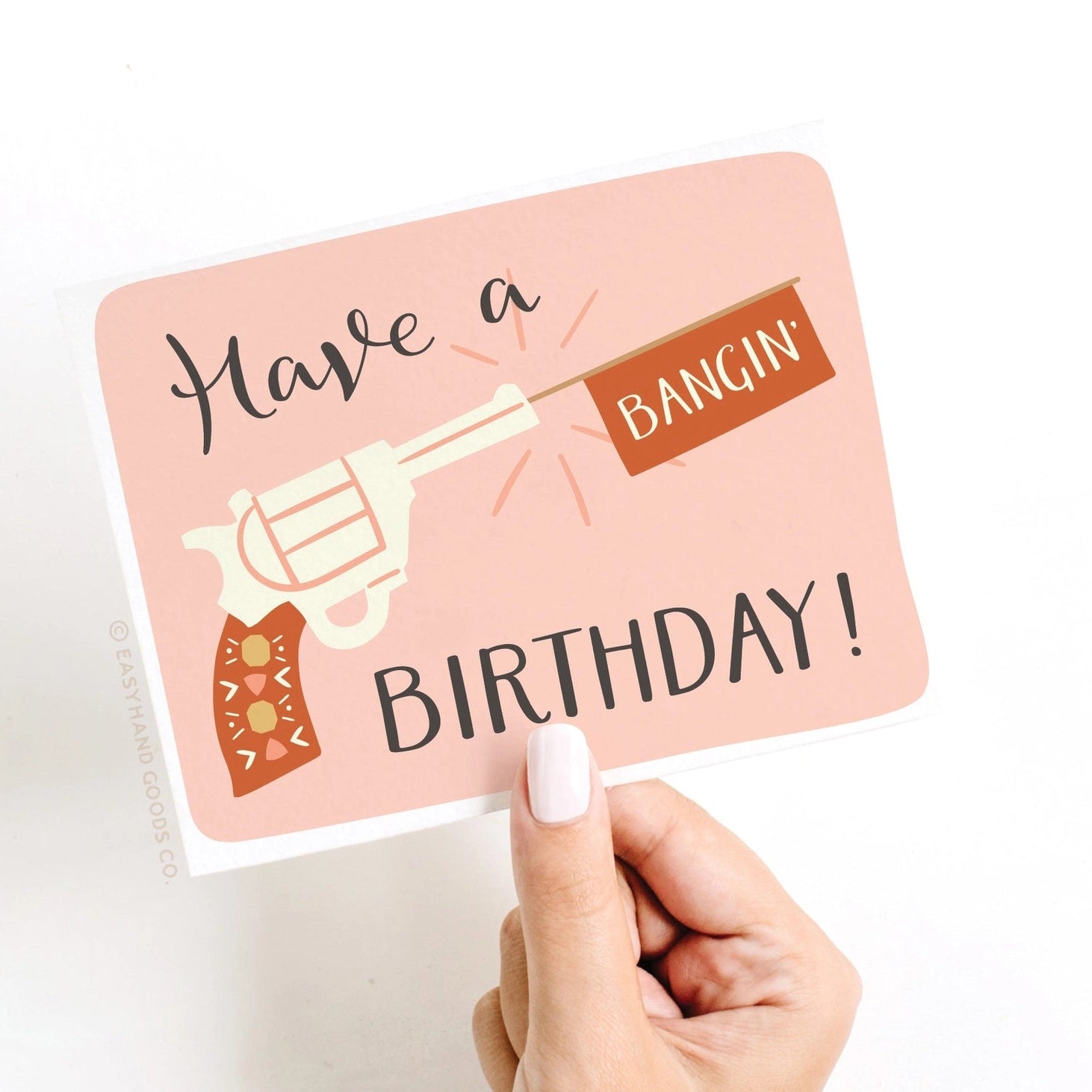 Hand holding a birthday card with a gun graphic and text on a pink background saying have a bangin birthday