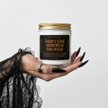 A hand with long black nails and a spider tattoo holds a glass jar candle against a light gray background. The candle has a gold metal lid, a white wax fill, and a black label with orange text that reads 'THERE'S SOME HORRORS IN THIS HOUSE'.