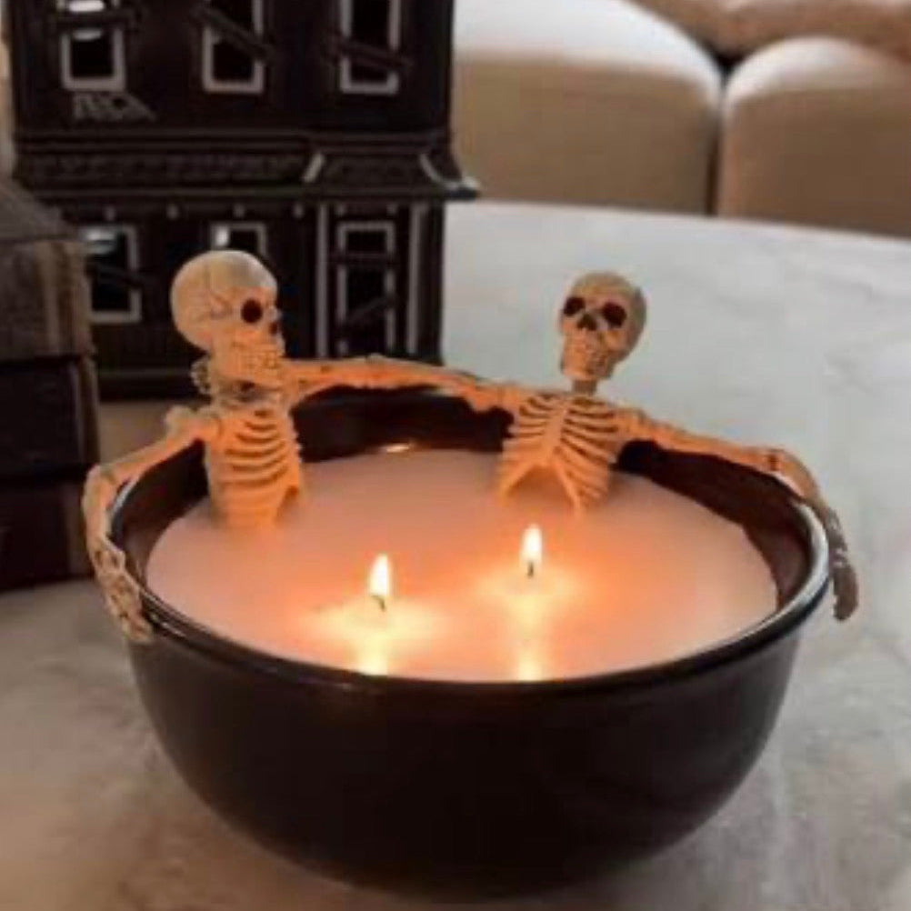 A black bowl candle with two lit wicks, decorated with two small skeleton figures sitting on the rim, set on a light-colored surface with a house-shaped ornament in the background.