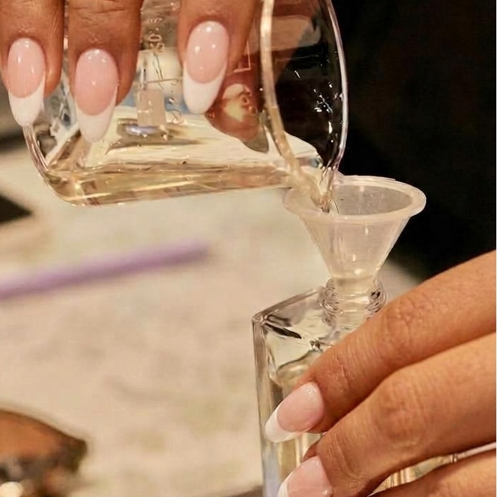 A close-up of hands with manicured nails pouring a clear liquid from a glass container into a small clear bottle, with a digital scale and cosmetic dropper bottles in the background.
