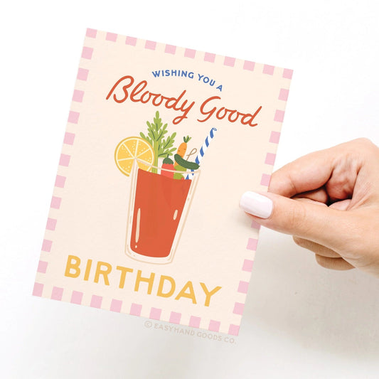 Hand holding a birthday card with a Bloody Mary illustration and text on a white background