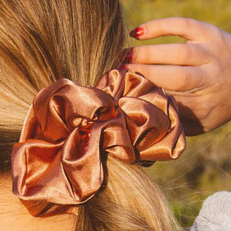 Person wearing a large, shiny bronze scrunchie on their hair with a blurred natural background.
