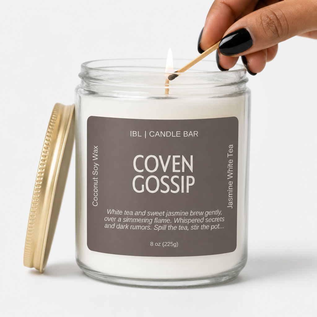 An 8 oz glass jar candle labeled 'Coven Gossip' by IBL Candle Bar, shown with its gold lid off and a hand lighting the candle with a wooden match. The candle has white wax and a dark brown label.