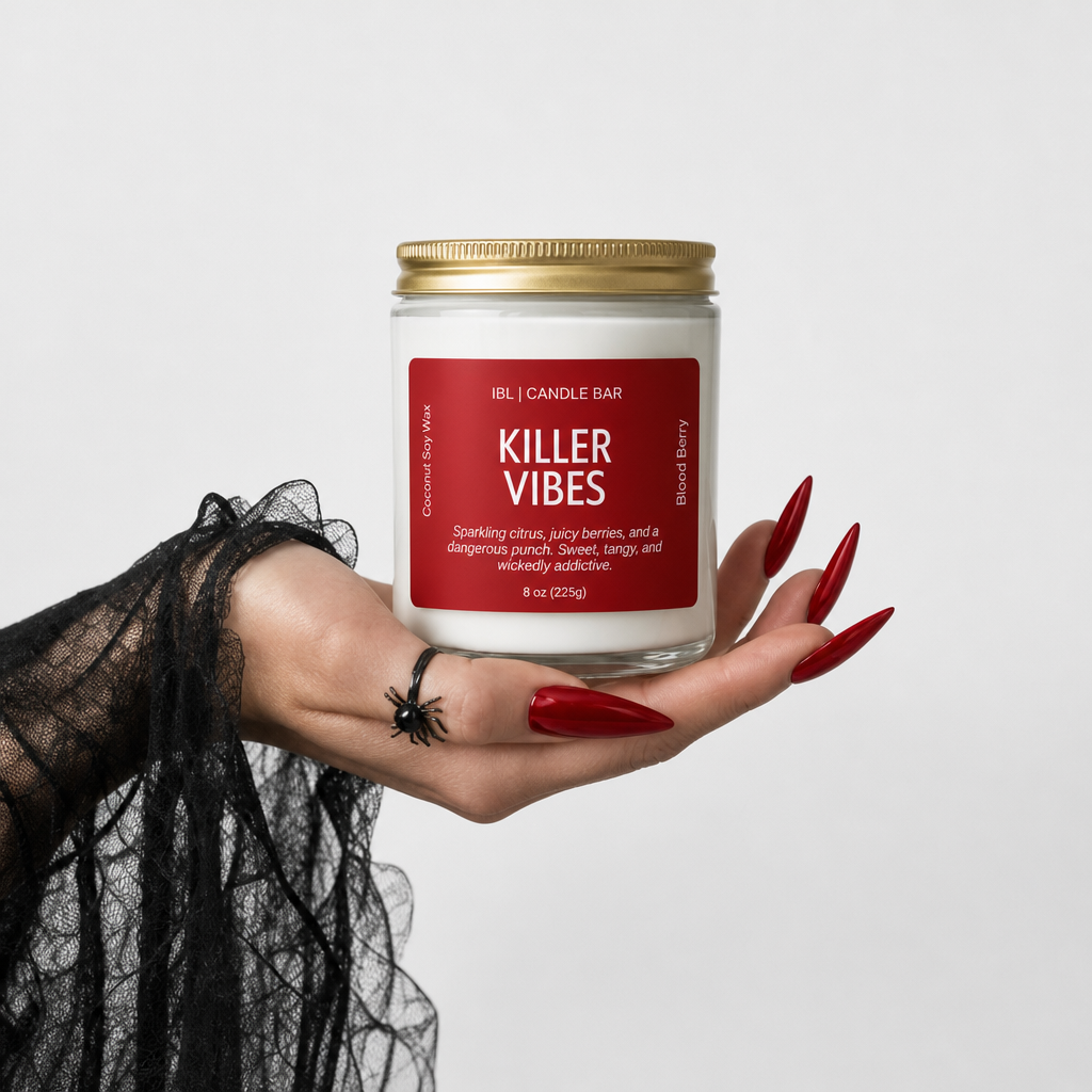 A hand with long red nails and a black spider tattoo holds a candle jar labeled 'Killer Vibes' by IBL Candle Bar. The candle is in a clear glass jar with white wax, a gold metal lid, and a red label.