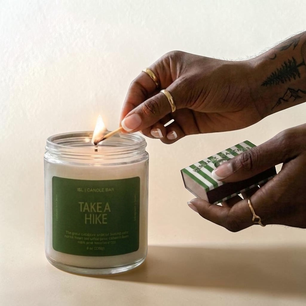 A glass jar candle labeled 'EL CANDLE BAY TAKE A HIKE' is lit with a wooden match held by a person's hand. The person also holds a green-and-white striped matchbox against a warm beige background.
