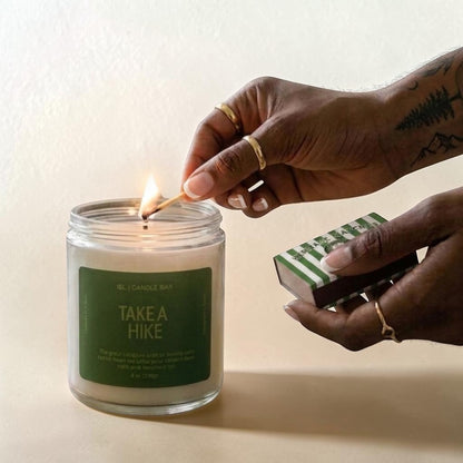 A glass jar candle labeled 'EL CANDLE BAY TAKE A HIKE' is lit with a wooden match held by a person's hand. The person also holds a green-and-white striped matchbox against a warm beige background.